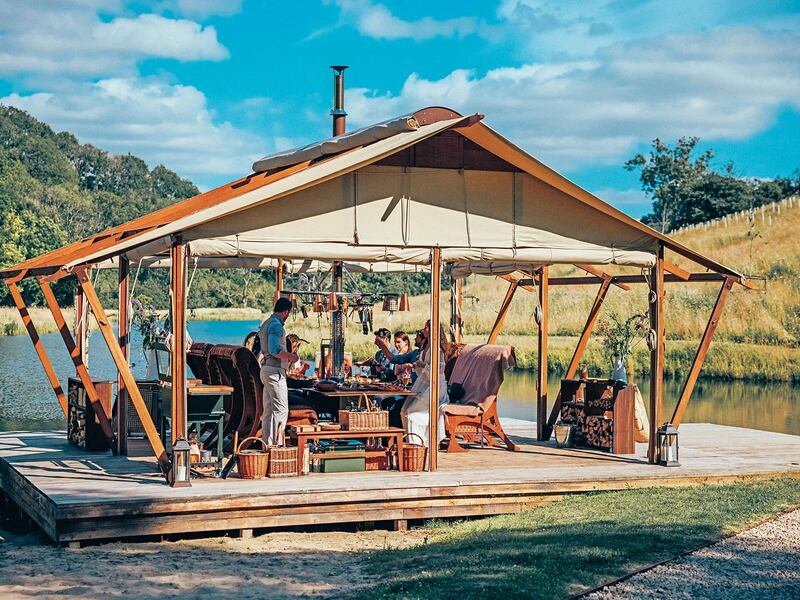 A floating platform in a lake backed by a hill covered in trees holds a pitched-roofed wooden structure with open sides and rolled up canvas that can be dropped down to create walls. It houses an outdoor kitchen, where a man is cooking.