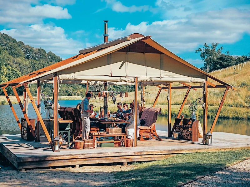 A floating platform in a lake backed by a hill covered in trees holds a pitched-roofed wooden structure with open sides and rolled up canvas that can be dropped down to create walls. It houses an outdoor kitchen, where a man is cooking.