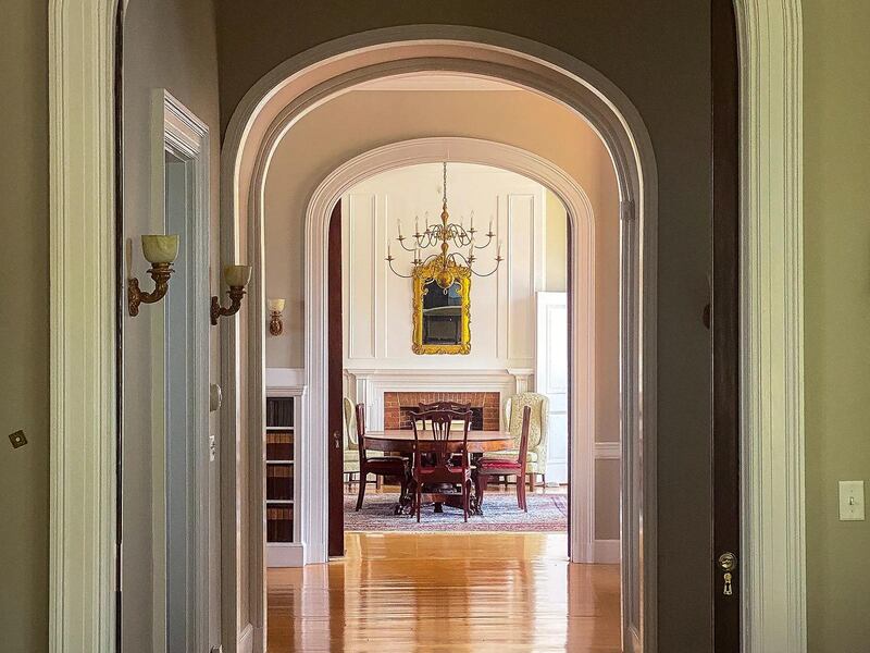 Arched entrances in a high-ceilinged hallway lead to a dranf room with white alls, a chandelier over a wooden table and chairs, and polished wooden floors