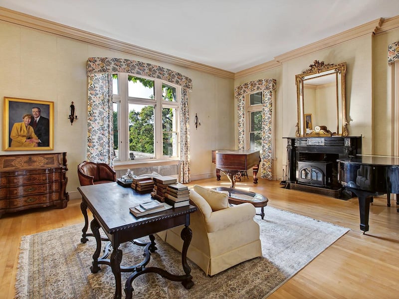 A bright living room with large windows, wooden floors and a feature fireplace with a mirror above it. A sofa faces a grand piano and has an antique table backing it, and a chest of drawers has a portrait painting of a couple on top of it.