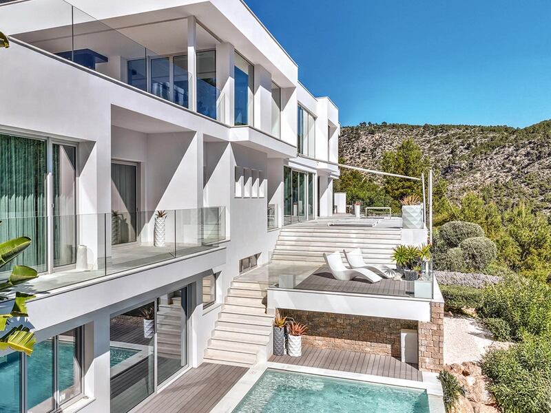 A rectangular swimming pool is on a terrace at the foot of a white-stone and glass villa, with a palm tree in the foreground and wooded mountain scenery in the background